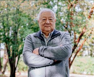 An elderly Asian man stands in front of some trees with his arms folded across his chest. 
